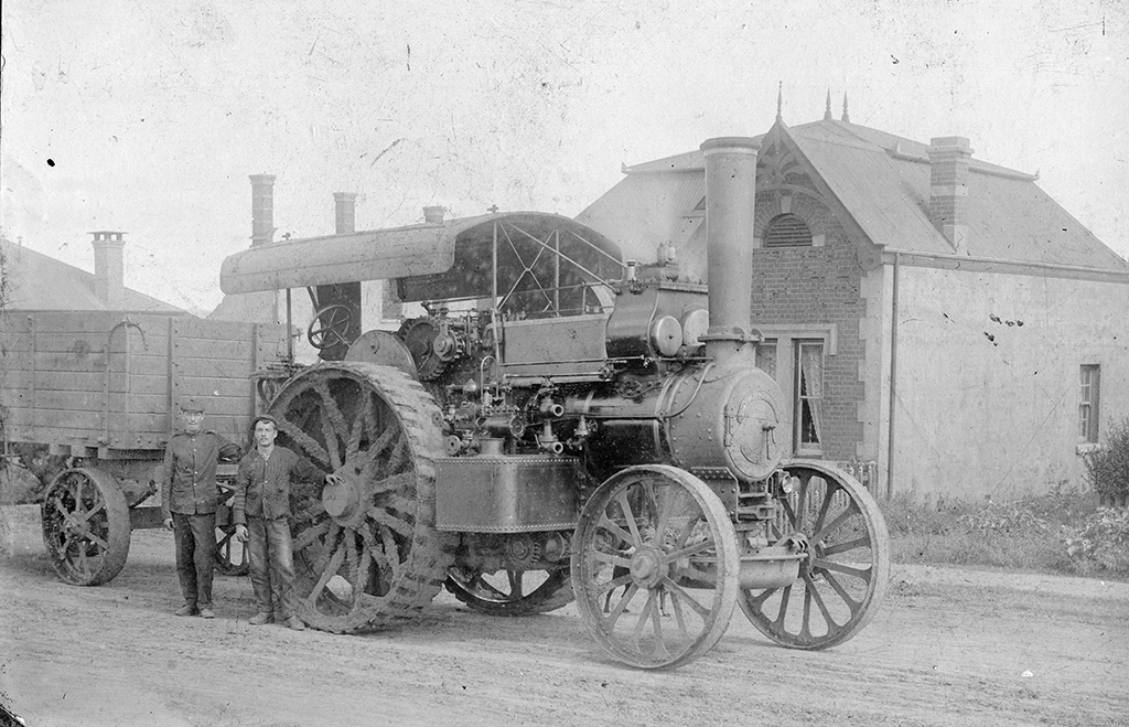 An unidentified Fowler B5 class road locomotive dating from the Boer War period supplied to the War Office. These engines were supplied to a very high specification and fitted with copper firebox’s, brass tubes, deep hind tanks, full width front tanks, and all the shafts were from Taylor s Gun steel all hardened in oil. Two men pose by the rear wheel looking towards the camera. It is attached to a Traction Wagon Trailer which is partially shown.