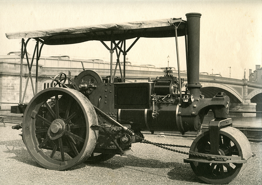 A photograph of an Aveling & Porter Steam Road Roller, a three quarter view of the offside. It is freshly painted and photographed  at the Strood, Rochester site, two arches of Rochester Bridge are shown in the background. 