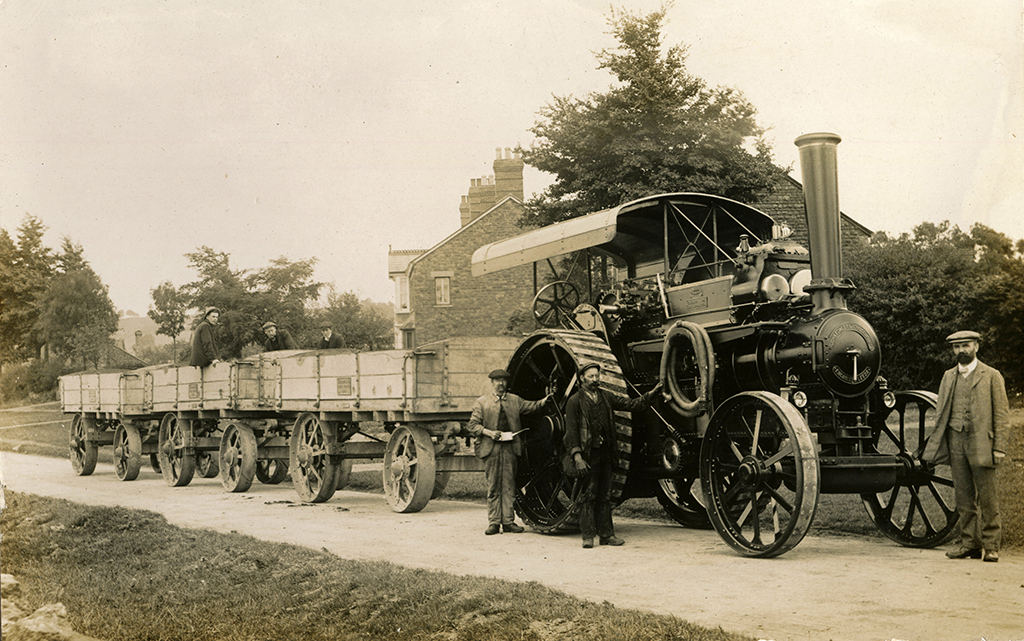 Fowler 7NHP road locomotive No. 12434 stands proudly hauling three traction wagons. Supplied new on 23rd September 1911 to James W. Harris & Son, Timber Merchants and Haulage Contractors, Hook Norton, Oxfordshire. Three men are seen standing next to the engine., three other men are sat in the second traction wagon. 