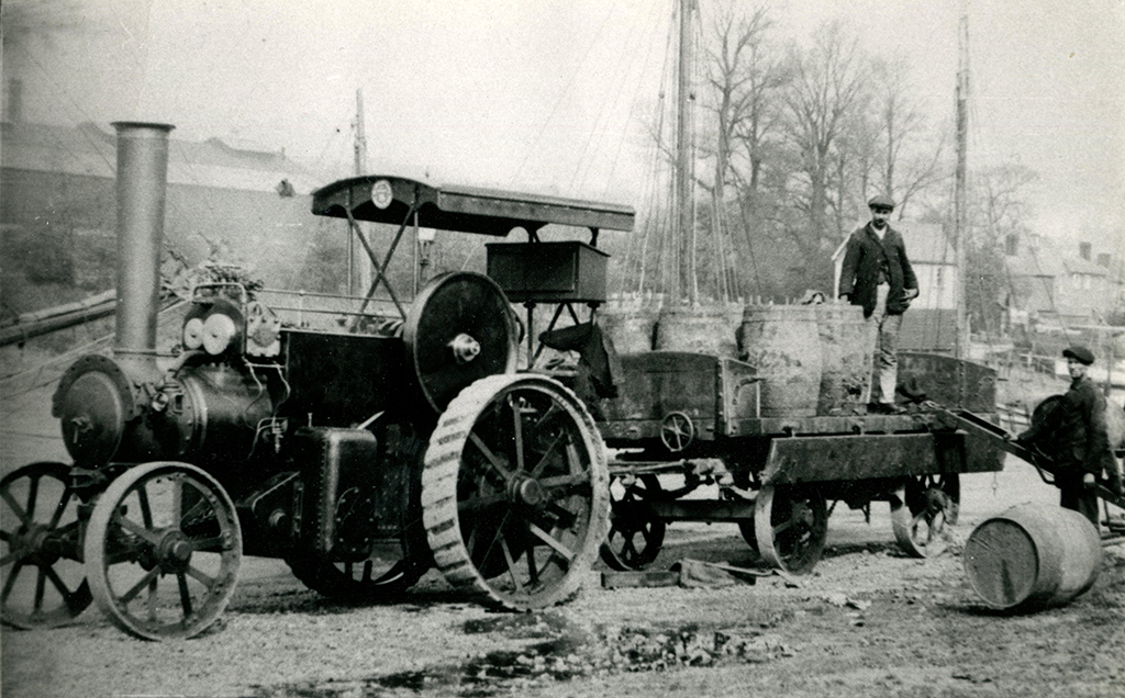 Isle of Wight Rural District Council’s Aveling & Porter convertible tractor No. 7577 of 1912 photographed at Newport, Isle of Wight. The photo shows the engine from the near-side facing left. It is towing a traction wagon with the near-side downcontainment board dropped to allow the loading or offloading of the load, which is of barrels. There is a man on the trailer and one on the ground., looking towards the reactor, One barrel has been unloaded.