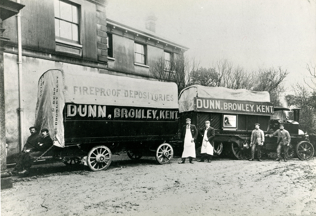 An early Foden Wagon  No. 642, registered D 593 on hire to H. C. Dunn & Sons, Bromley, Kent on removal from Chislehurst Mineral Water Co. The photograph shows the wagon facing away from the camera with its removal trailer. Both showing the sign written branding of the company. Both the wagon and trailer have canvas tops. Six staff are included, four standing in front of the Wagon and two are sat on the hinged trailer flap, which is close to the ground. 