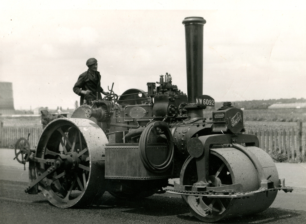 A 1964 black-and-white photo of a Fowler ‘T3’ steam roller (No. 15970) working on the A4 in Reading. The off-side view shows the machine facing right, with a driver on the footplate. A hose is coiled neatly between the rolls, a brass plate sits on the motion cover, and the rear roll features a scarifier.